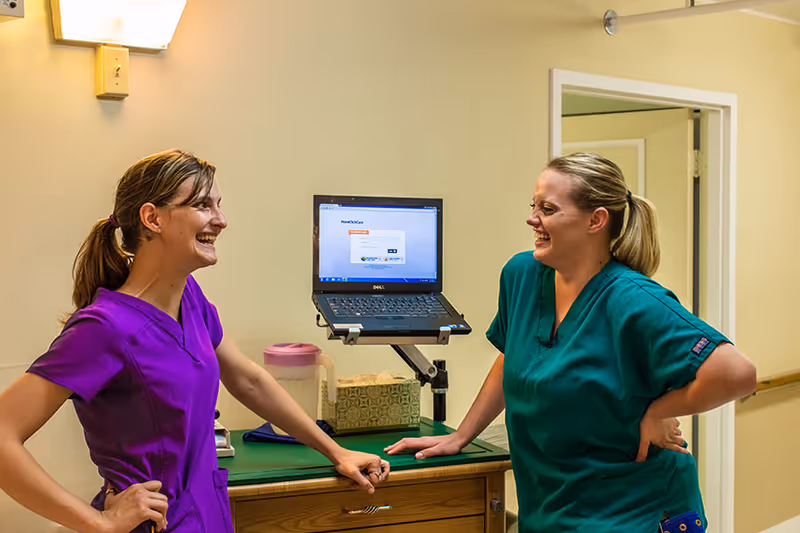 Two healthcare workers in scrubs, one in purple and one in green, smiling and talking in a hallway with a laptop on a stand and a tissue box on a counter behind them.