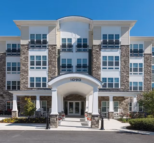 Four-story senior living building with a curved entrance, stone accents, and multiple balconies.