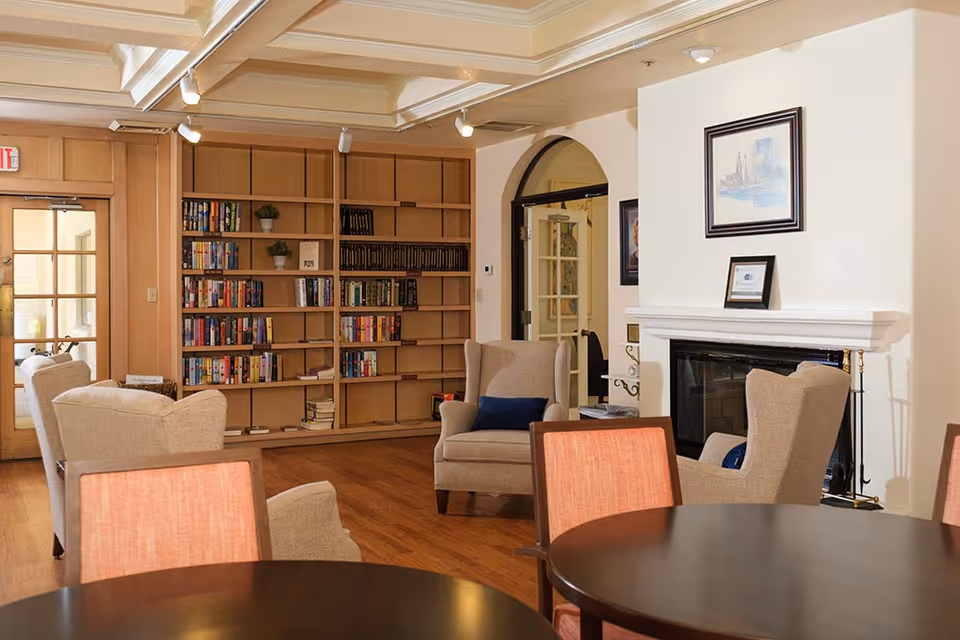 Cozy common room with bookshelves, armchairs, round tables, and a fireplace.