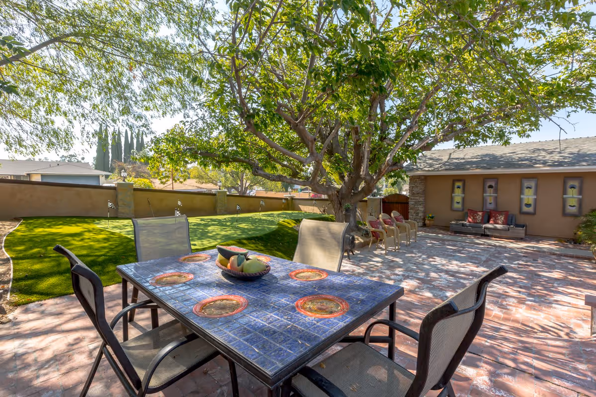 Outdoor patio area with a blue tiled table and four chairs under a large tree. In the background, there is a putting green with small flags and a seating area with multiple chairs and a couch against a tan wall with decorative wall art.