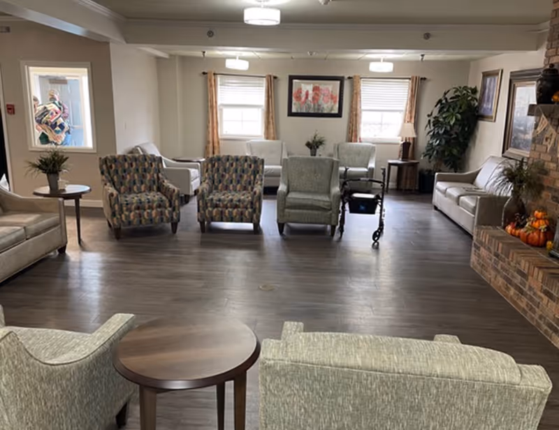 A spacious living room area in a senior living facility with multiple armchairs and sofas arranged around a central space. There is a brick fireplace on the right decorated with small pumpkins and a plant. Two windows with curtains and a framed floral picture are on the far wall. A walker is positioned near one of the chairs. The room has wooden flooring and ceiling lights.