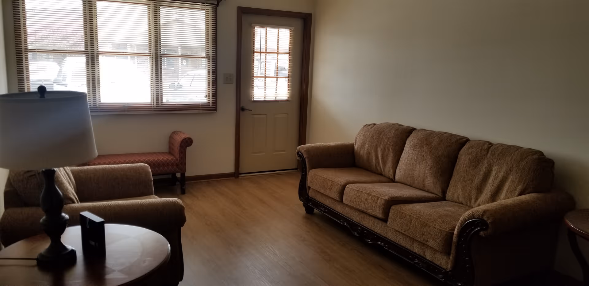 A cozy living room with a brown upholstered sofa and armchair, a wooden side table with a lamp, a small red bench under a window with blinds, and a door with a window panel. The floor is wooden and the walls are light-colored.