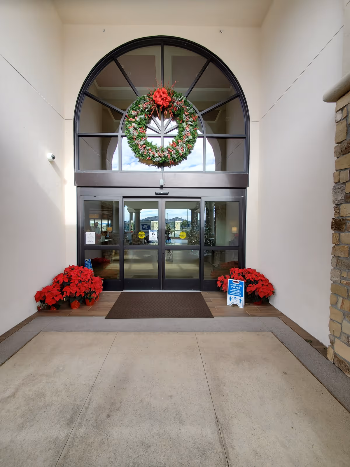 Entrance to a building with large glass double doors under a tall arched window decorated with a large green wreath with a red bow. There are red poinsettia plants on either side of the entrance and a small sign on the right side. The walls are light-colored and there is a stone column on the right.