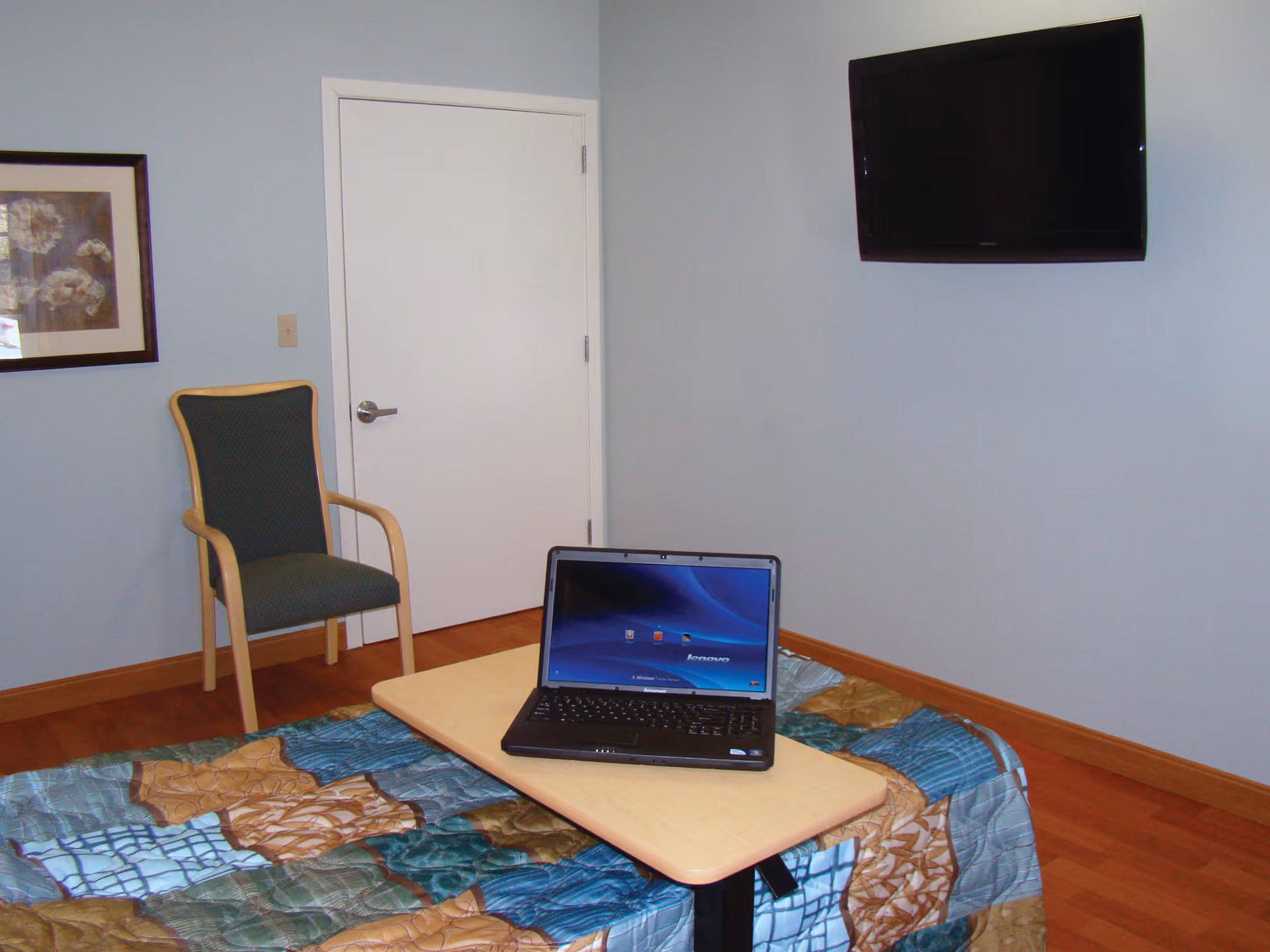 A small room with a bed covered by a colorful quilt, a wooden chair with green upholstery, a closed white door, a wall-mounted flat screen TV, and a laptop placed on a small table over the bed.
