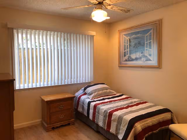 A small bedroom with a single bed covered in a striped bedspread in shades of red, black, gray, and white. Next to the bed is a wooden nightstand with two drawers. A large window with vertical blinds is on the left wall, and a framed picture of an open window looking out to a beach scene hangs on the wall above the bed. The ceiling has a fan with a light fixture.