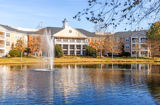 Three-story senior living building seen across a pond with a central water fountain and surrounding trees.