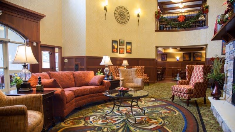 A cozy senior living facility common area with a curved red sofa, patterned armchairs, a round glass coffee table on a colorful patterned rug, and warm lighting from table lamps. The room features wood paneling, a stone fireplace decorated with holiday garlands, and a high ceiling with a balcony above.