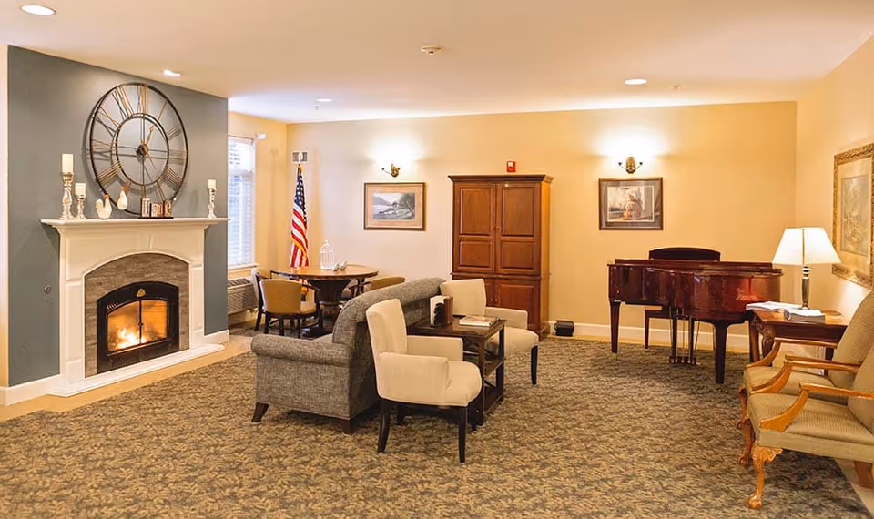 A cozy senior living community lounge with a lit fireplace, a large wall clock above the mantel, comfortable seating including armchairs and a sofa, a wooden cabinet, a grand piano, framed artwork on the walls, an American flag near a window, and warm lighting from wall sconces and a table lamp.