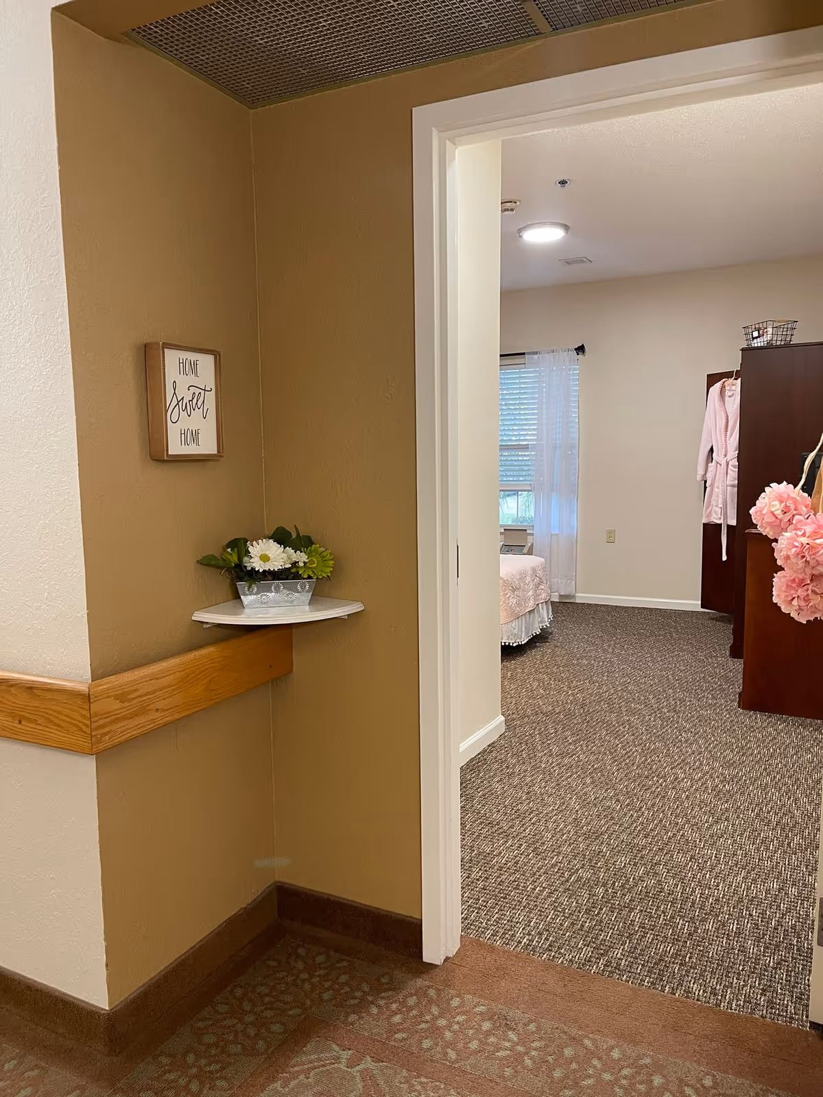 Hallway entry into a resident bedroom in an assisted living facility, showing a 'Home Sweet Home' sign, a small shelf with flowers, and a partial view of a bed and wardrobe.