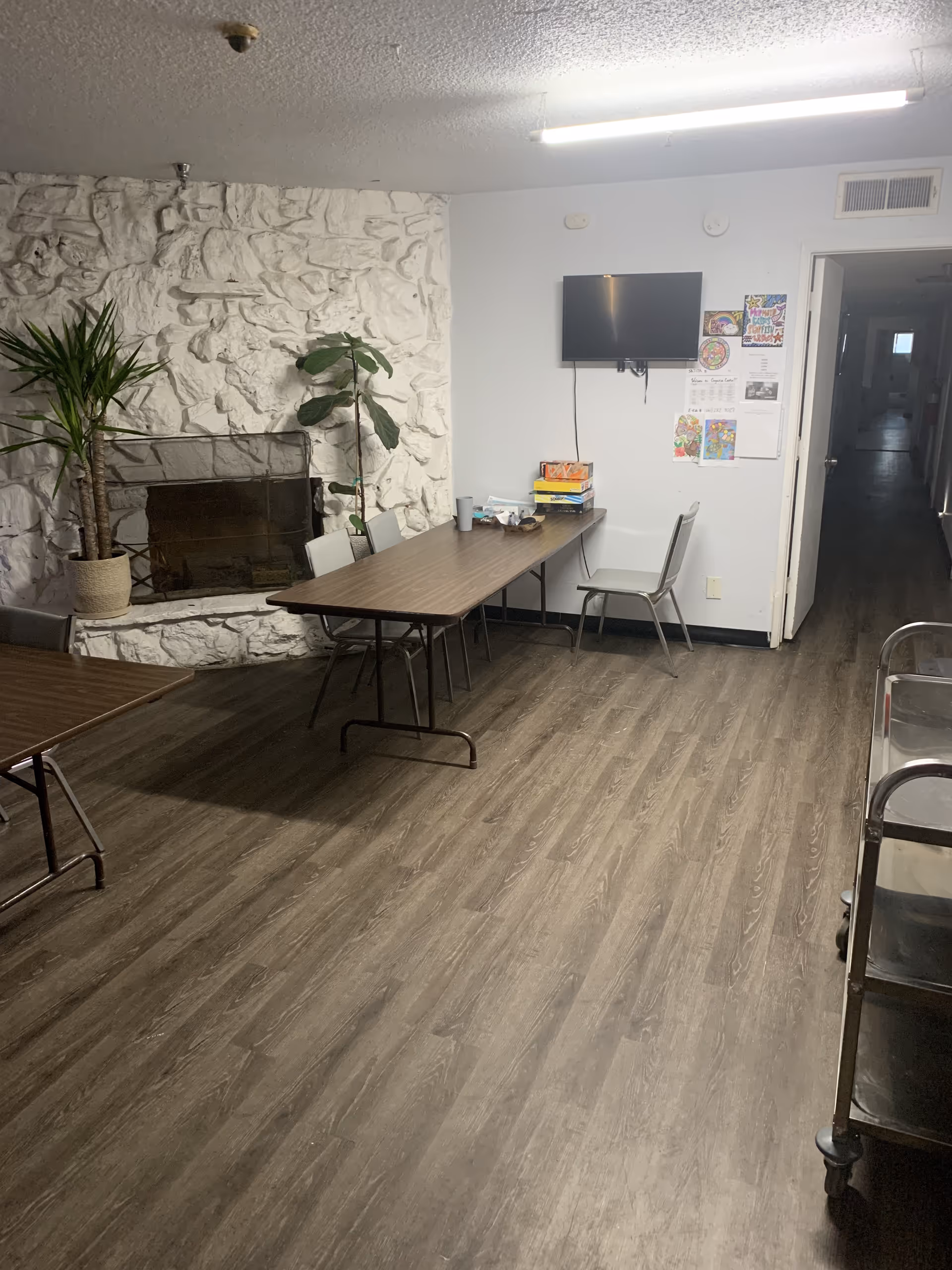 Interior room with wood flooring, a white textured stone fireplace, two potted plants, a long table with chairs, a wall-mounted TV, and a hallway leading to other rooms. There are some papers and board games on the table.
