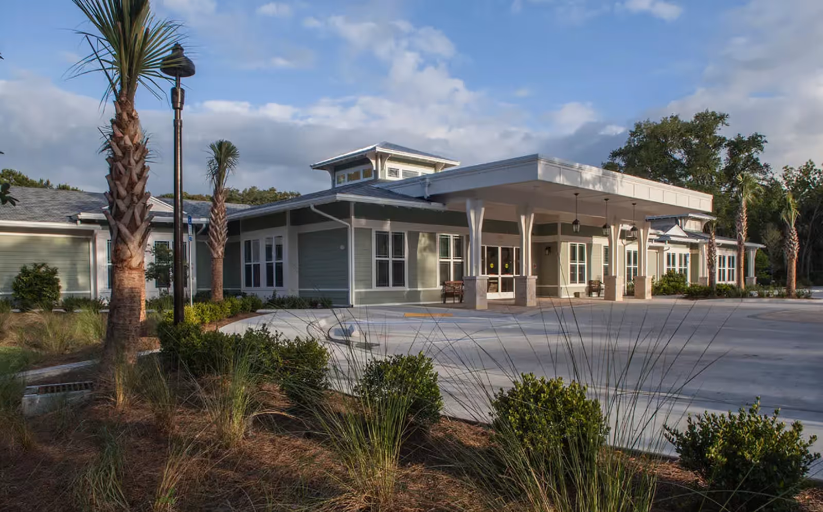 Exterior view of Mount Pleasant Gardens facility showing a single-story building with a covered entrance, surrounded by palm trees and landscaped bushes under a partly cloudy sky.