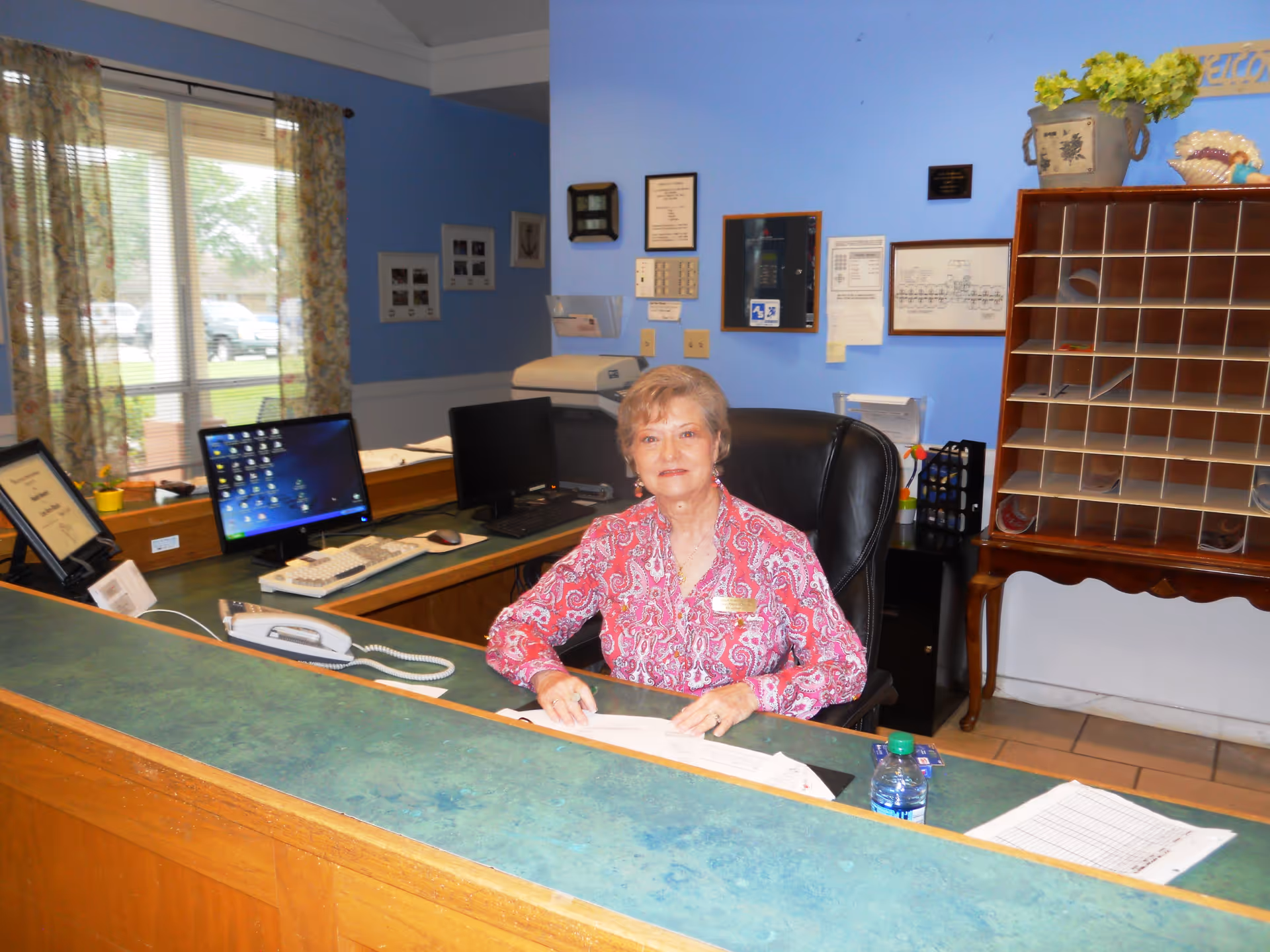 An elderly woman sitting behind a reception desk in an office area with a computer, telephone, and paperwork. The walls are painted blue and decorated with framed certificates and a mail organizer. A window with floral curtains is visible in the background.