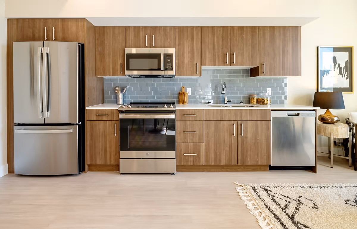 Modern kitchen with wooden cabinets, stainless steel refrigerator, oven with stovetop, microwave, dishwasher, and a sink. The backsplash is made of light blue subway tiles. There is a small wooden side table with a lamp and framed artwork on the wall to the right.