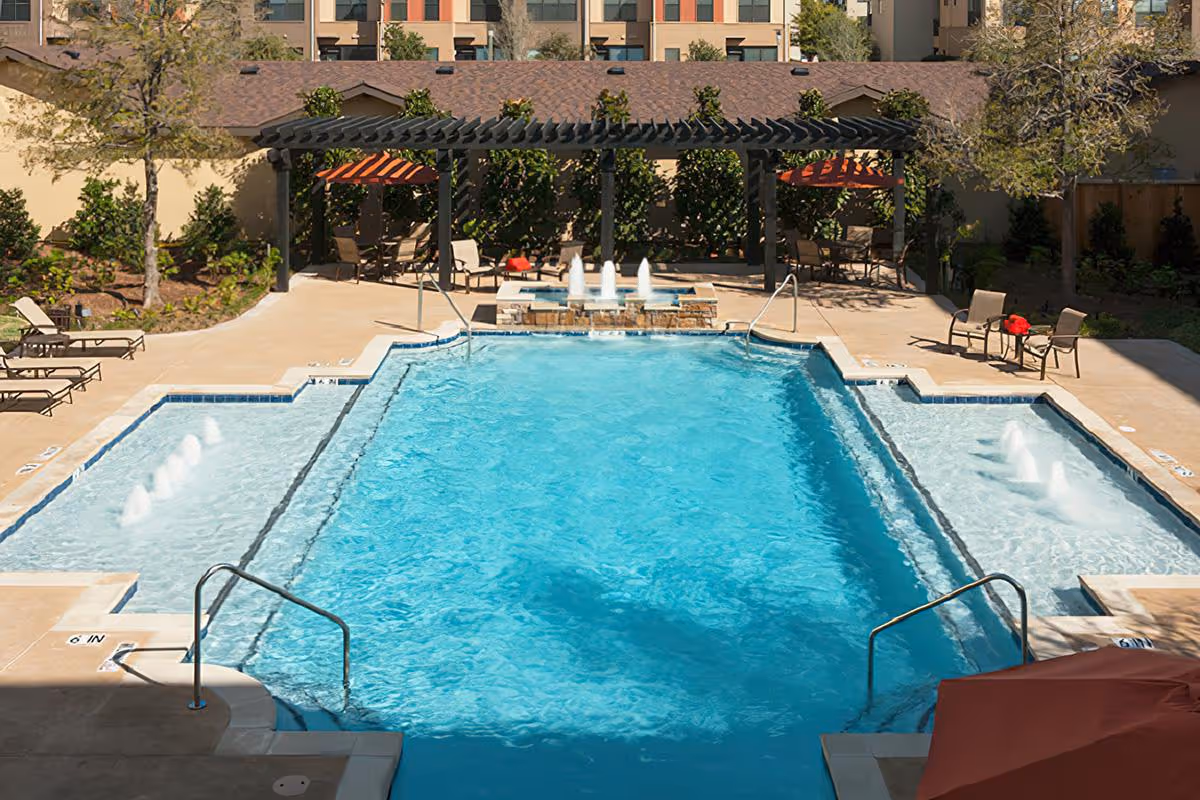 Outdoor swimming pool with shallow wading areas, fountains, lounge chairs, and a shaded pergola at a residential facility.