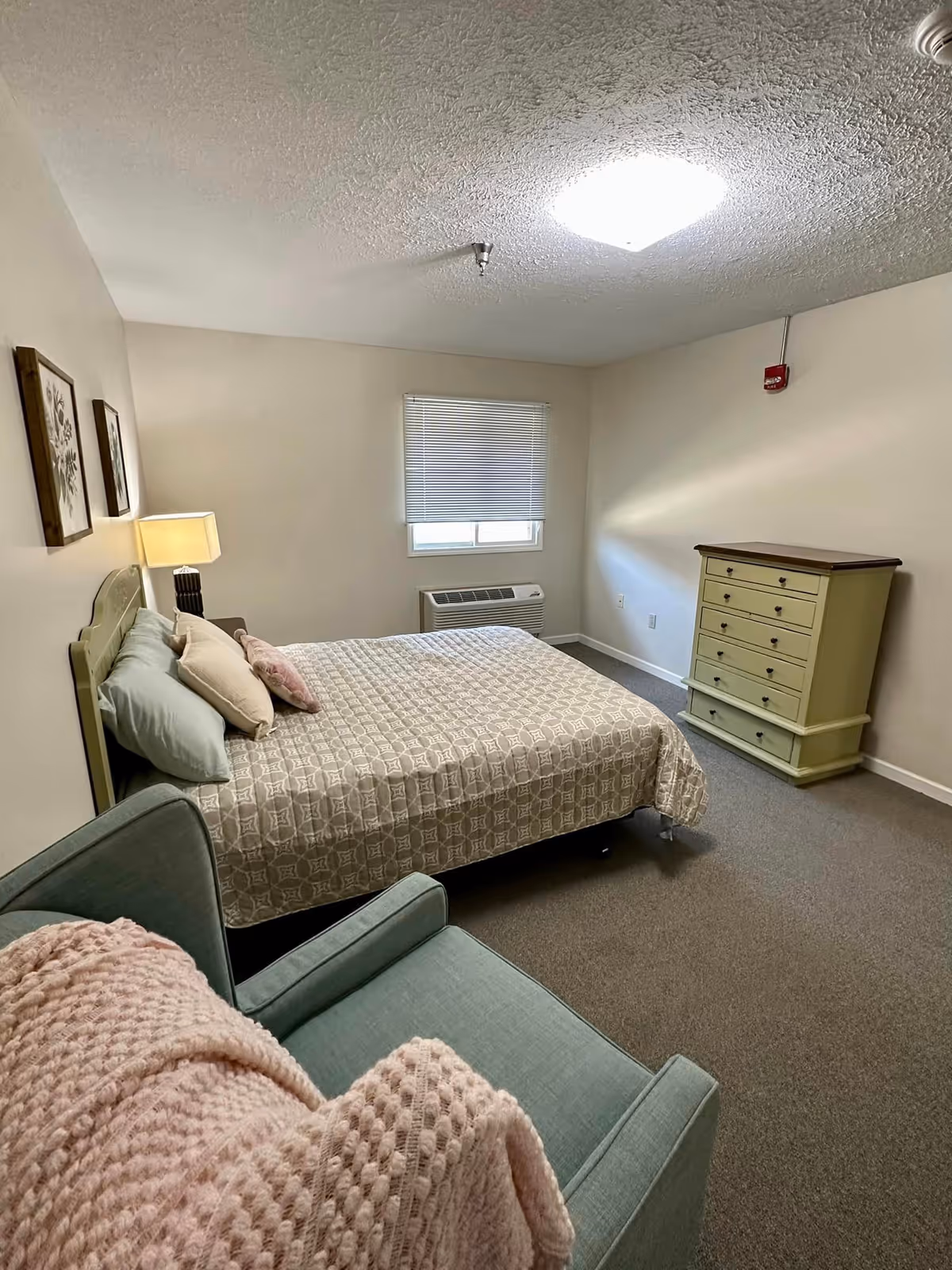 A simple bedroom with a bed covered in a patterned beige bedspread and several pillows. There is a green armchair with a pink knitted blanket draped over it in the foreground. On the right side of the room is a green dresser with multiple drawers. A window with closed blinds is above a wall-mounted air conditioning unit. The walls are plain and light-colored, and the ceiling has a textured finish with a bright ceiling light.