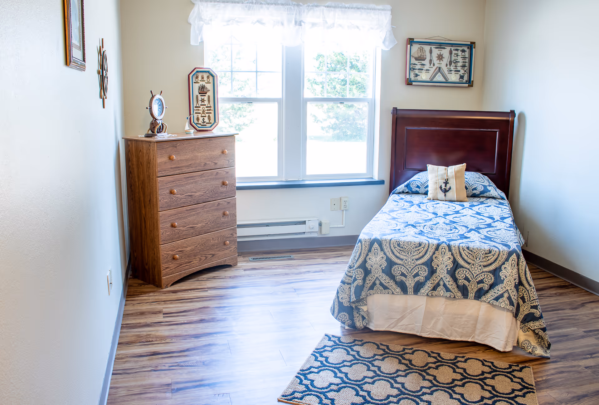 A small, neatly arranged bedroom with a single bed featuring a blue and white patterned bedspread and a pillow with an anchor design. There is a wooden dresser with four drawers to the left of the bed, and two framed nautical-themed pictures on the walls. A window with white curtains lets in natural light, and a patterned rug is placed on the wooden floor near the foot of the bed.