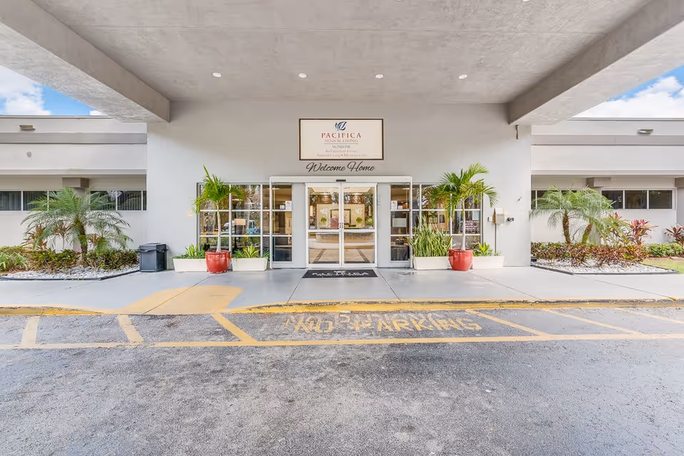 Entrance of a senior living facility with automatic sliding glass doors, potted plants on either side, and a sign above that reads 'Pacifica Senior Living Sunrise Independent Living Assisted Living Memory Care' with the words 'Welcome Home' underneath. There are landscaped areas with palm trees and shrubs on both sides of the entrance and a 'No Parking' zone marked on the pavement in front.