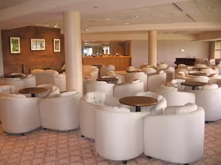 A spacious lounge area with multiple round tables surrounded by white cushioned chairs. The room features beige carpeting, brick accent walls with framed artwork, and several support columns. There is a piano visible in the background near large windows letting in natural light.