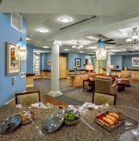 Interior view of a senior living facility dining and common area with a granite countertop in the foreground displaying covered dishes, a bowl of salad, and a tray of cooked chicken. The room features blue walls, decorative columns, hanging pendant lights, ceiling fans, framed artwork, and comfortable upholstered chairs arranged around small tables.