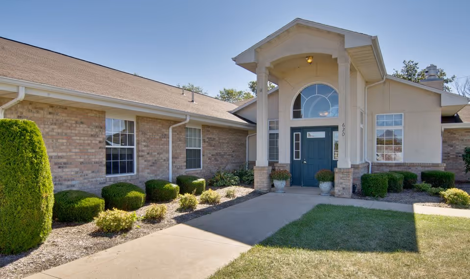 Exterior view of a single-story brick building with a covered entrance featuring two white columns and a blue door. The building is surrounded by neatly trimmed bushes and a well-maintained lawn under a clear blue sky.