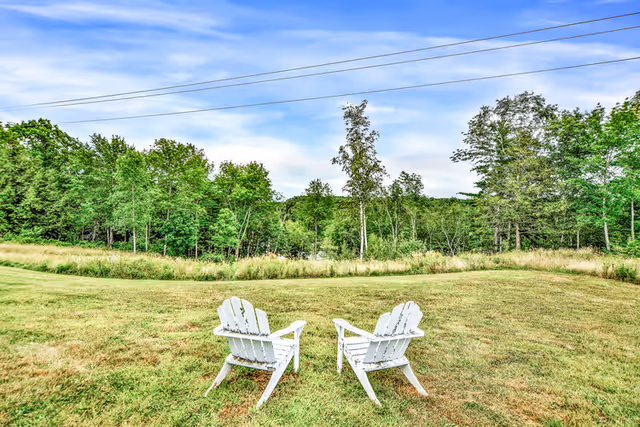 Two white Adirondack chairs on a grassy lawn facing a treeline under a blue sky.