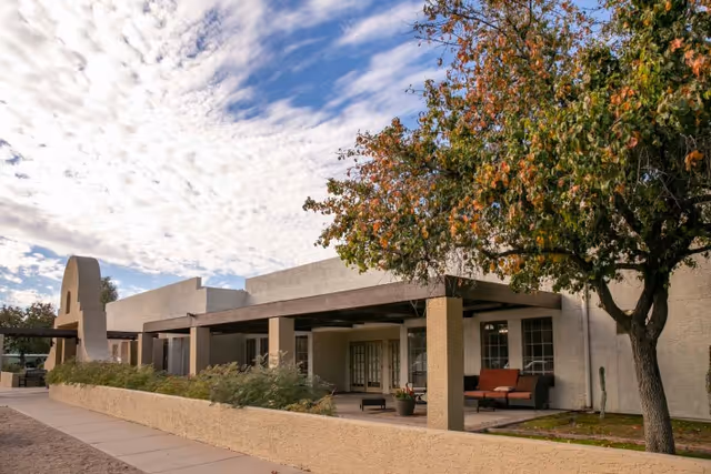 Covered patio seating and entrance of a stucco rehabilitation building with a tree and partly cloudy sky.