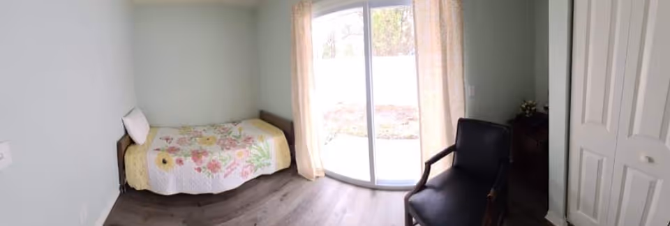 A small bedroom with a single bed covered with a floral quilt, a white pillow, a black armchair, a wooden nightstand with a small plant, and a sliding glass door with light curtains leading outside. The room has light-colored walls and wood flooring.