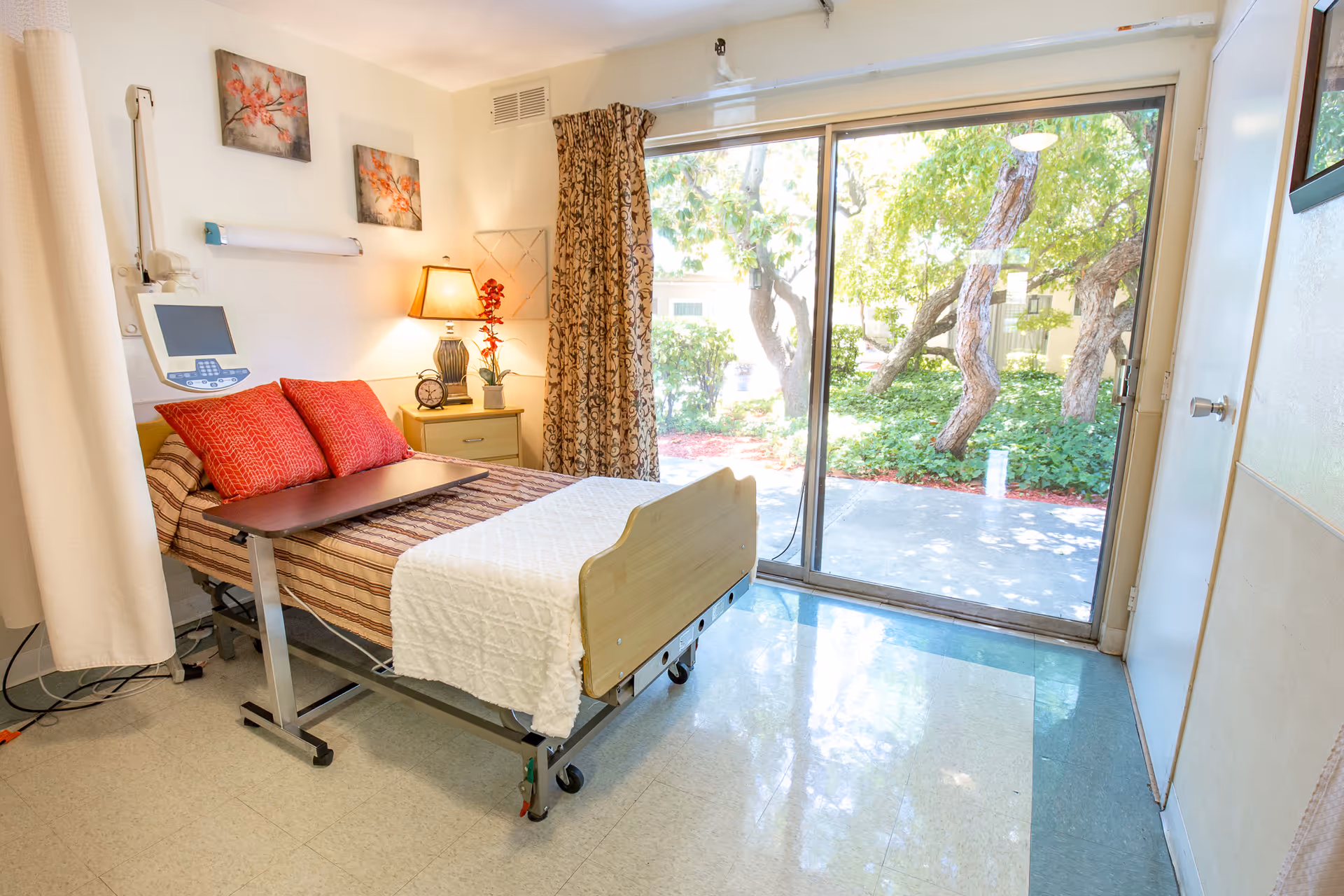 A bright and clean patient room with a hospital bed featuring red pillows and a white blanket. There is a bedside table with a lamp, clock, and a small plant. The room has large sliding glass doors that open to a garden area with trees and greenery outside. The floor is tiled, and there are two floral paintings on the wall above the bed.