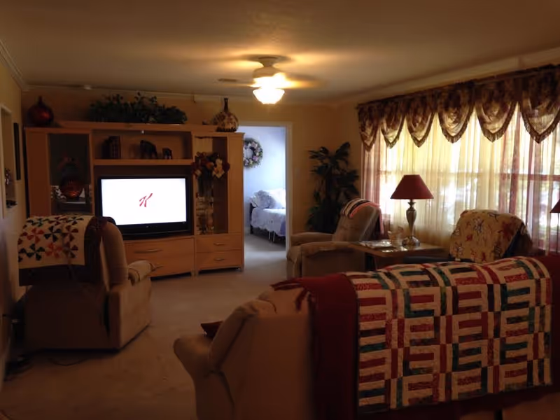 A cozy living room with beige armchairs and a sofa draped with colorful quilts. A wooden entertainment center holds a television displaying a red 'K' logo. The room is softly lit by a ceiling light and natural light coming through large windows with sheer curtains and valances. A side table with a lamp and decorative plants add to the homey atmosphere. In the background, a bedroom with a bed and floral wreath on the wall is visible through an open doorway.