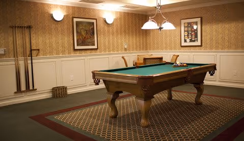 A billiards table in a carpeted game room with cue racks, framed artwork, and overhead lighting.