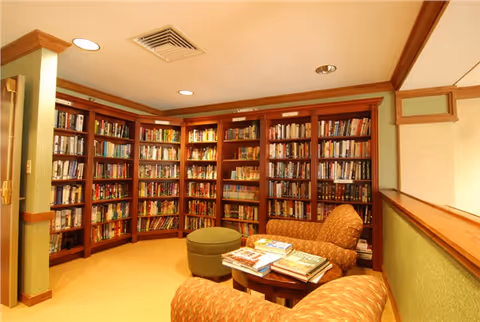 A cozy library room with wooden bookshelves filled with books lining the walls. There are two patterned armchairs and a small round table with several books stacked on it. The room has warm lighting and a beige carpeted floor.