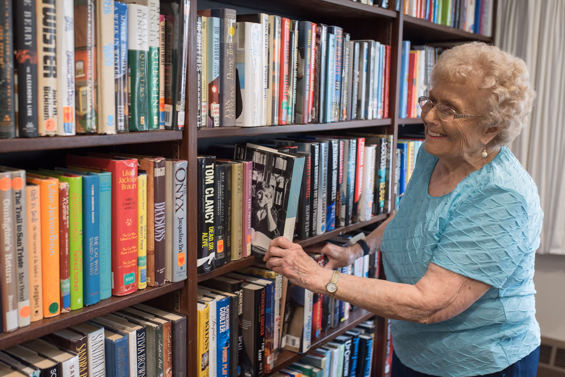 An elderly woman with curly hair and glasses, wearing a light blue textured blouse, is smiling as she selects a book from a wooden bookshelf filled with various books in a well-lit room.