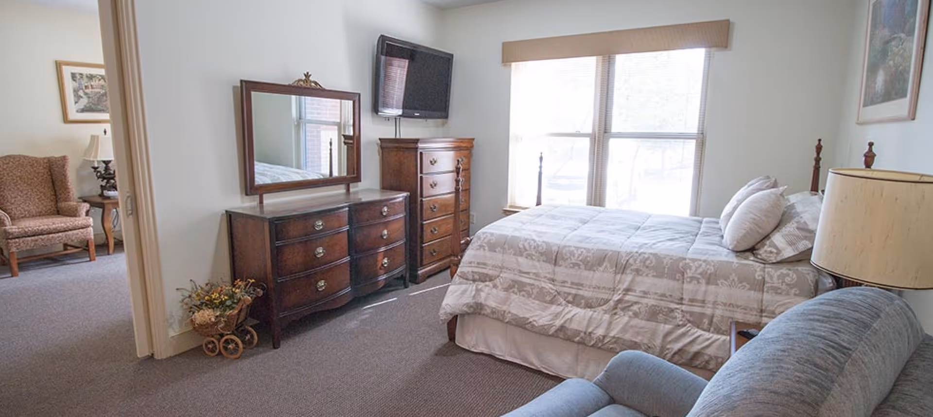 A bedroom with a bed covered in a beige patterned comforter, two pillows, a wooden dresser with a mirror, a tall chest of drawers, a wall-mounted TV, a blue armchair, and a lamp on a side table. There is a window with blinds letting in natural light and an open doorway leading to another room with an upholstered armchair and a side table with a lamp.