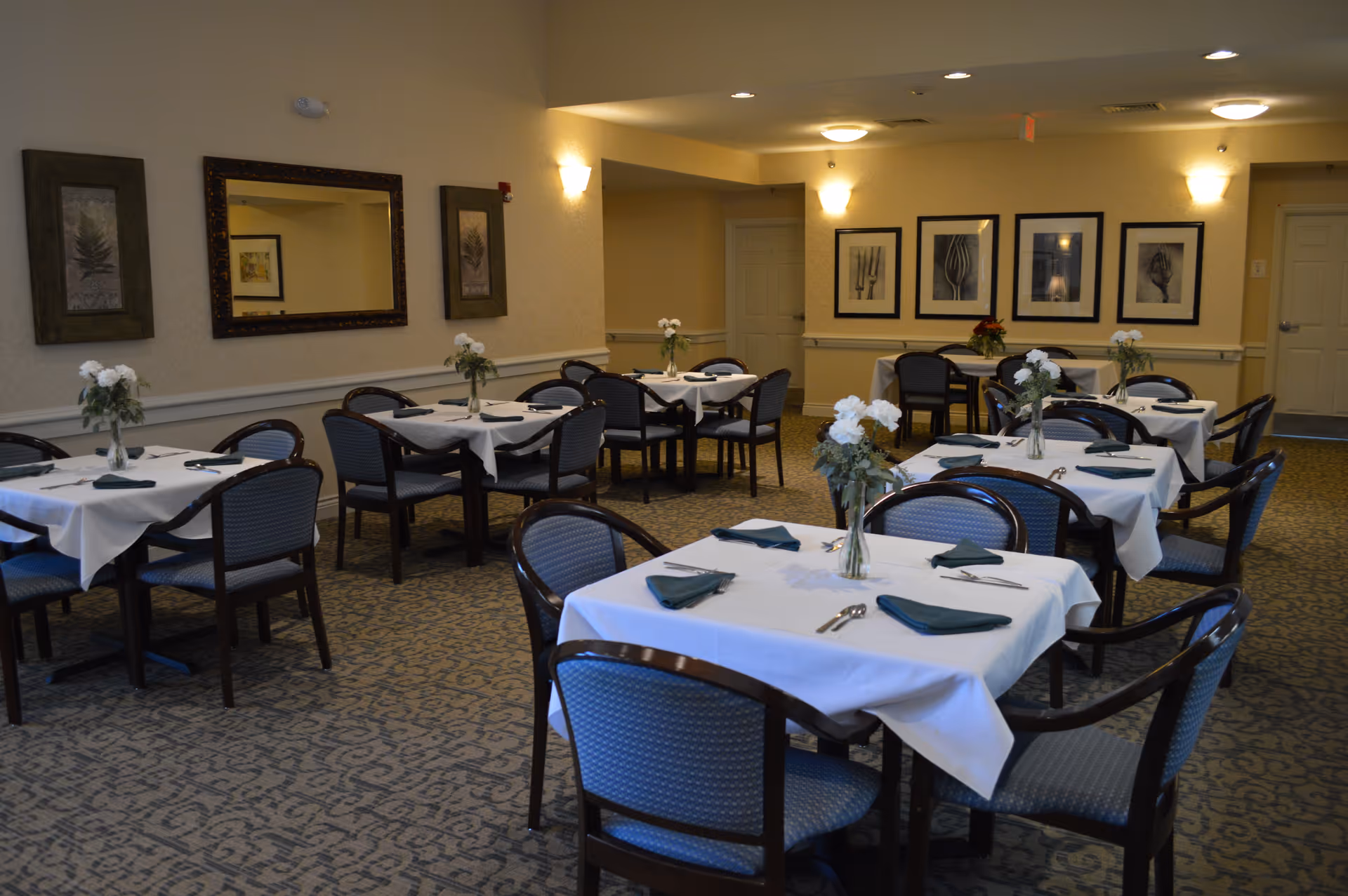 Dining room with several tables covered in white tablecloths, green napkins, chairs, and flower centerpieces.