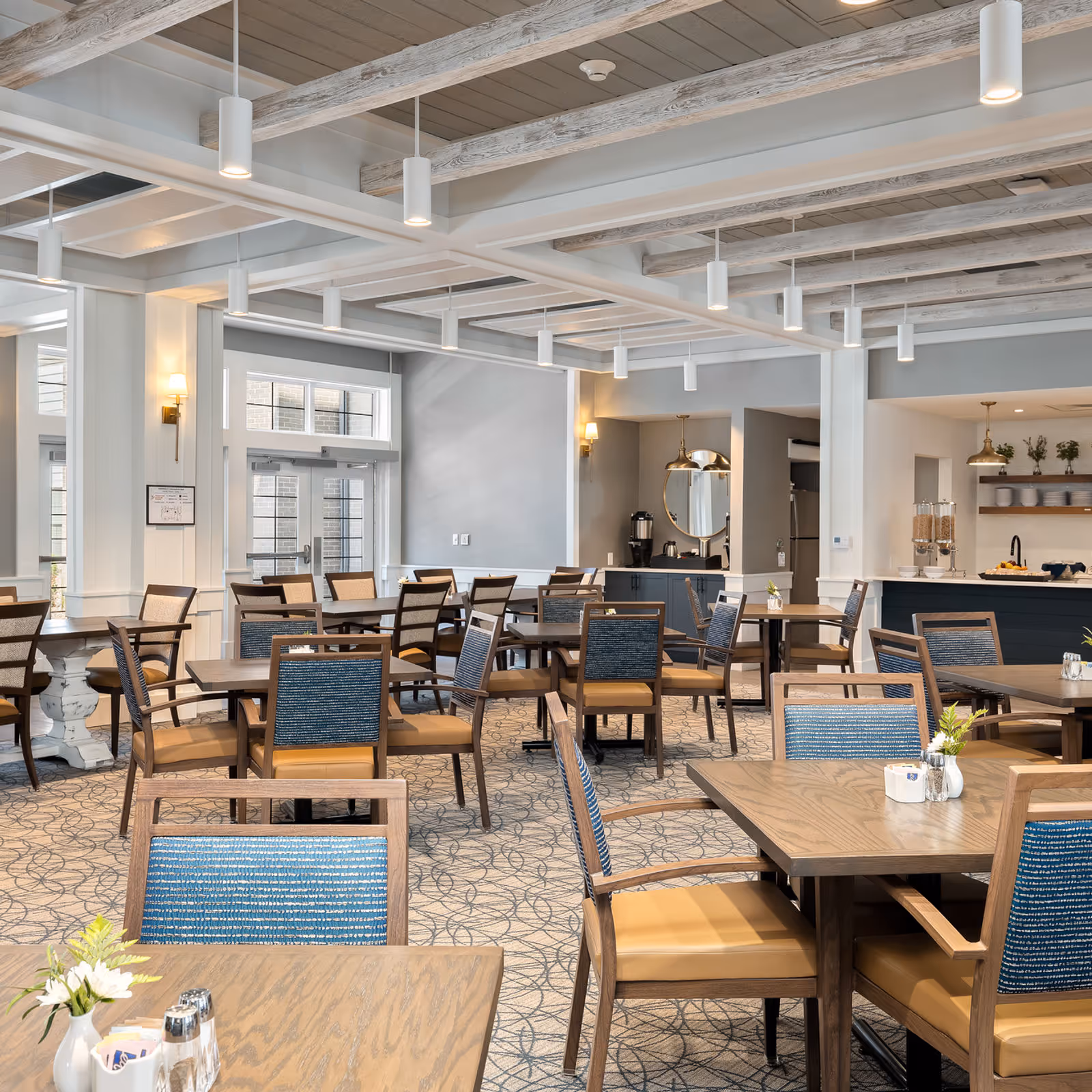 A spacious and well-lit dining room with multiple wooden tables and chairs featuring blue and tan upholstery. The room has a patterned carpet, white walls with wainscoting, and a ceiling with exposed wooden beams and hanging pendant lights. In the background, there is a counter area with cereal dispensers, plates, and a coffee station.