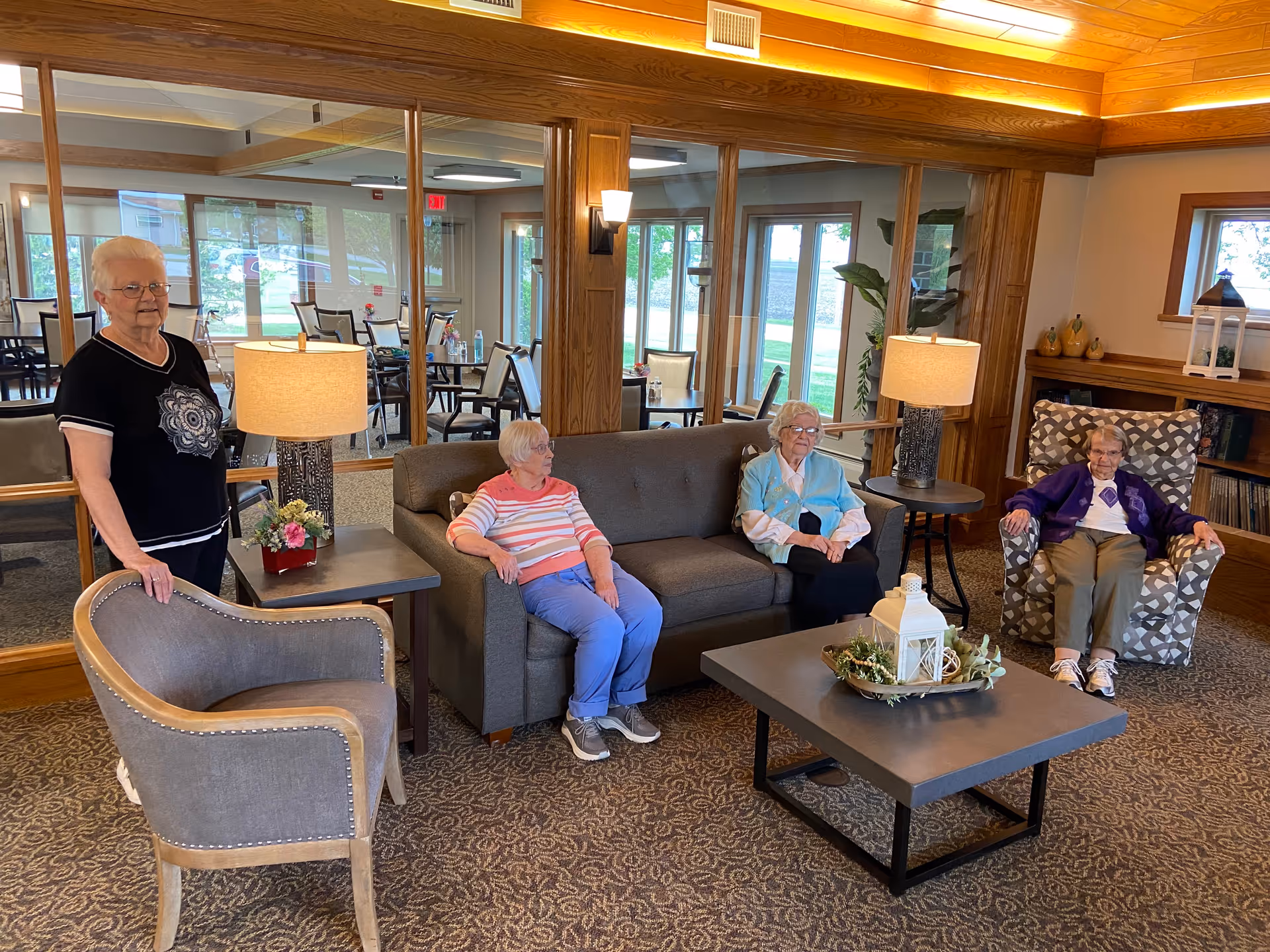 Four seniors in a communal living room sitting on a sofa and armchairs around a coffee table with lamps and decor.