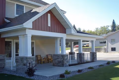 Exterior view of a residential building with a covered porch supported by white columns and stone bases. Two wooden chairs are placed on the porch. The building has red and beige siding with a sloped roof. There is a driveway and another building visible in the background along with some trees.