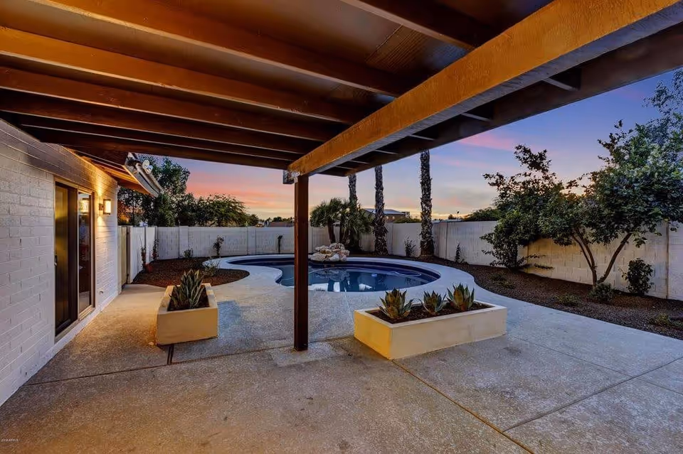 Covered patio area with wooden beams overlooking a kidney-shaped swimming pool surrounded by a concrete deck, with planters containing succulents and a white brick wall enclosing the backyard. Trees and shrubs are planted along the perimeter, and the sky shows a colorful sunset.