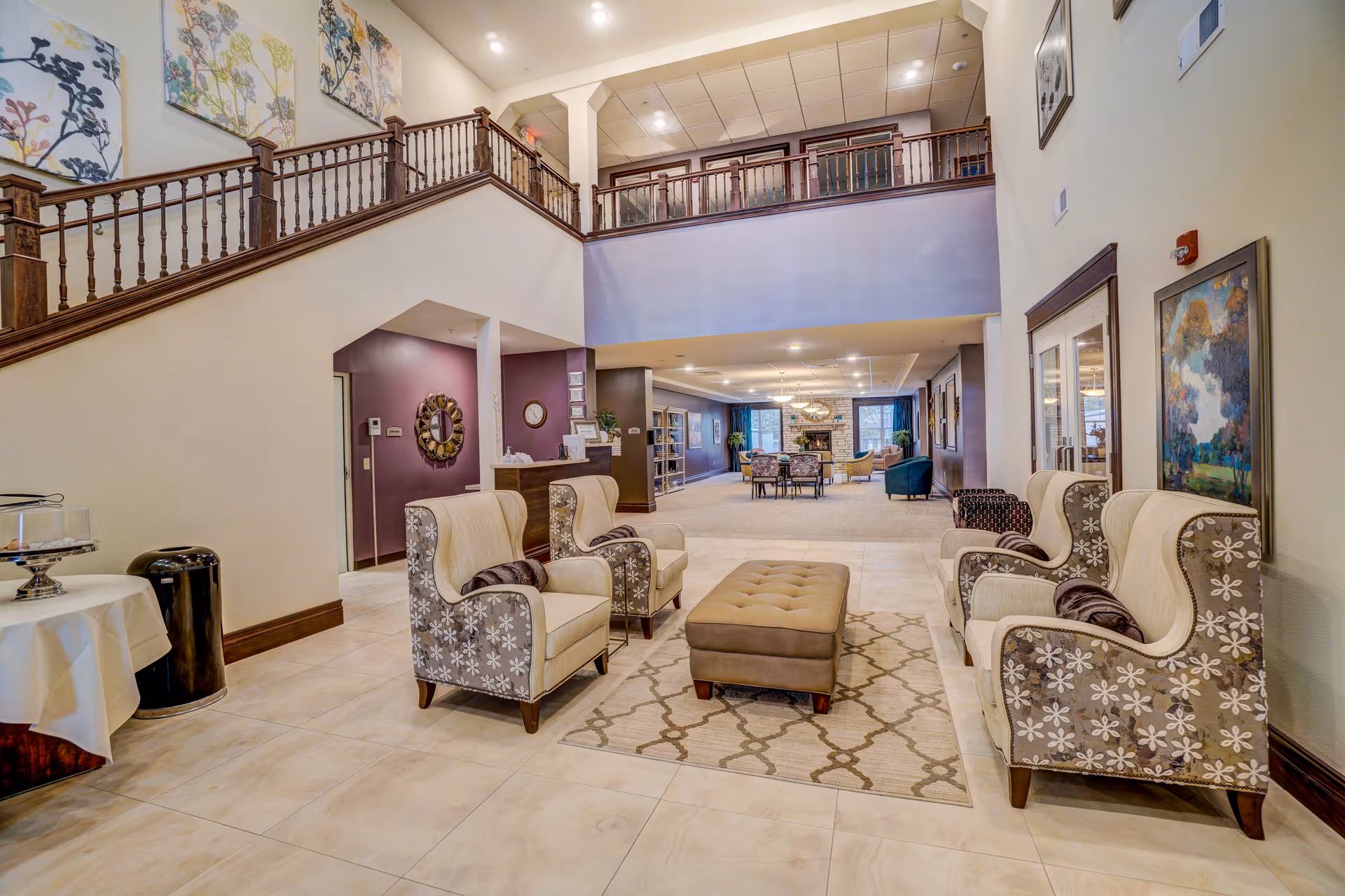 Spacious senior living facility common area with four patterned armchairs arranged around a rectangular ottoman on a decorative rug. The room features a high ceiling with a balcony railing above, light-colored tiled floor, and walls adorned with framed artwork. In the background, there is a dining area with tables and chairs near large windows and a stone fireplace.