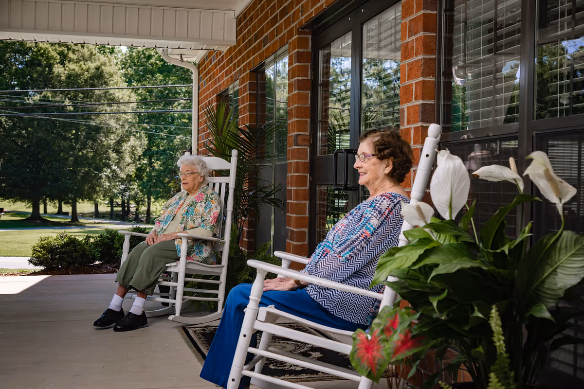 Two elderly women sitting in white rocking chairs on a covered brick porch overlooking a green lawn.