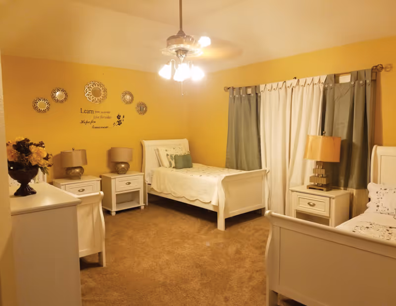 Yellow-walled bedroom with two white beds, nightstands, lamps, a ceiling fan, and curtained window.