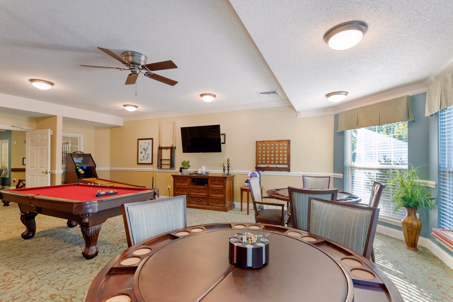 A well-lit game room with a red felt pool table, a poker table with chairs, a wall-mounted TV, and a rack holding pool cues. There is a large window with blinds and a potted plant near it. The room has beige walls with white trim and a ceiling fan with wooden blades.