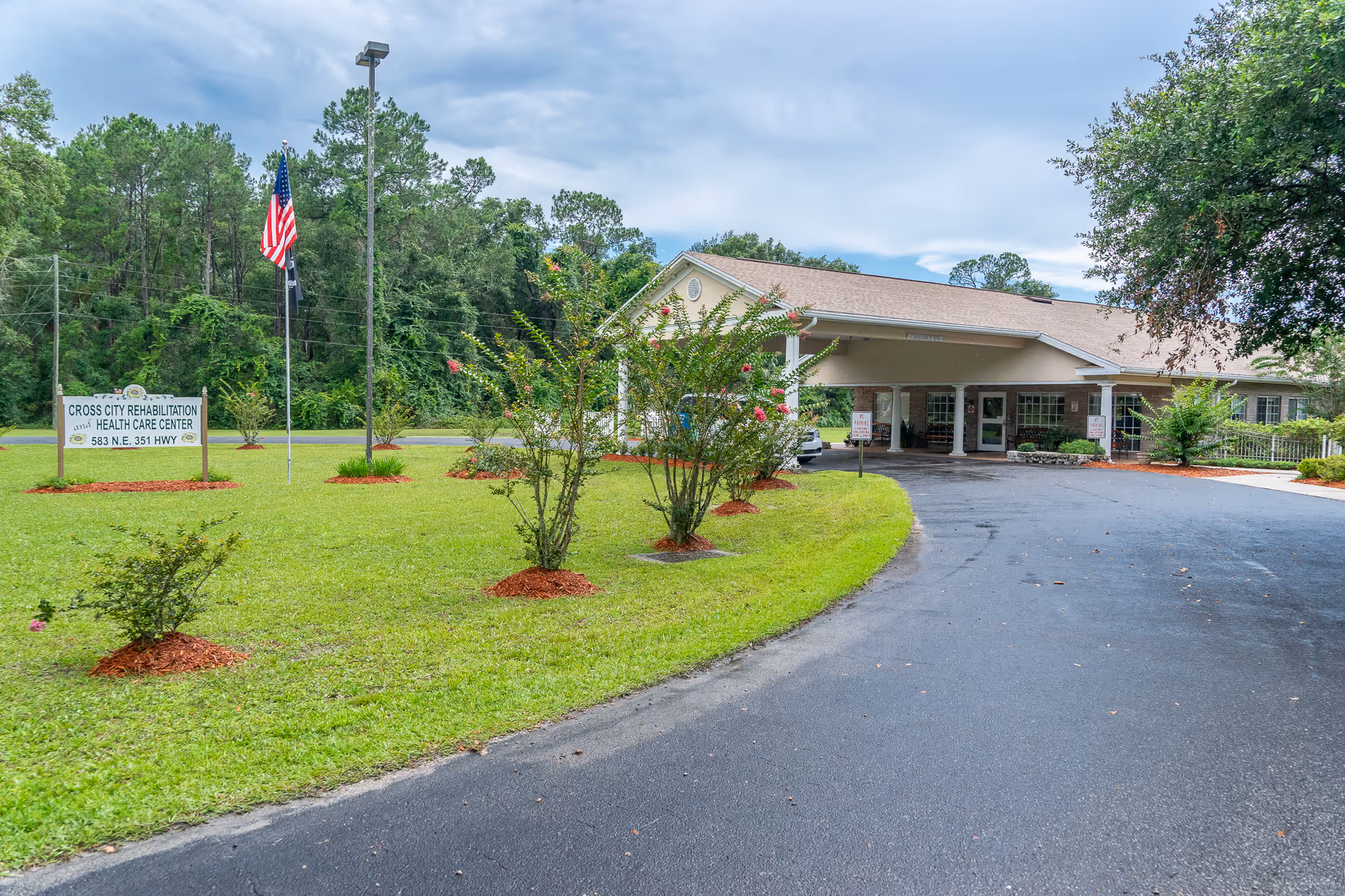 Exterior view of Cross City Rehabilitation Health Care Center showing a single-story building with a covered entrance, a curved driveway, a green lawn with small trees and bushes, an American flag on a flagpole, and a sign displaying the facility's name and address.