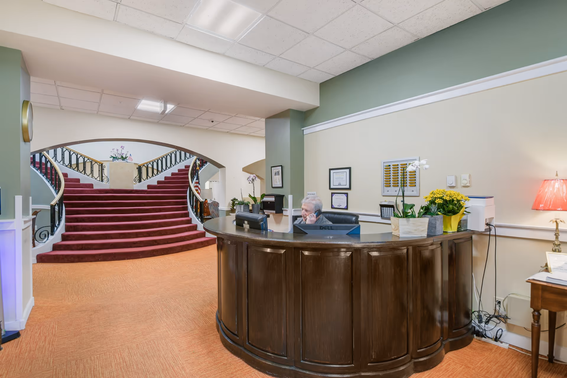 Reception area of Drum Hill Senior Living Community featuring a curved wooden front desk with a receptionist on the phone. Behind the desk are framed certificates on the wall, potted plants, and a printer. In the background, a wide staircase with red carpet and black railings splits into two directions under an archway.