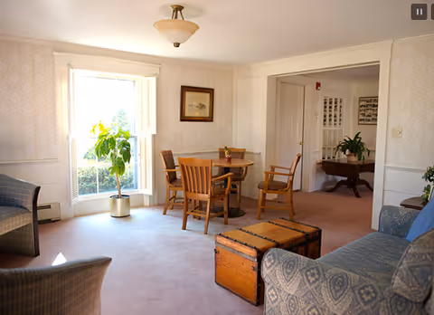 A bright living room area with a round wooden table and four chairs in the center. There is a large window with white shutters letting in natural light, and a potted plant near the window. The room has light-colored walls with subtle wallpaper and carpeted flooring. A patterned sofa and an old wooden trunk are visible in the foreground, with an open doorway leading to another room with more furniture and plants.