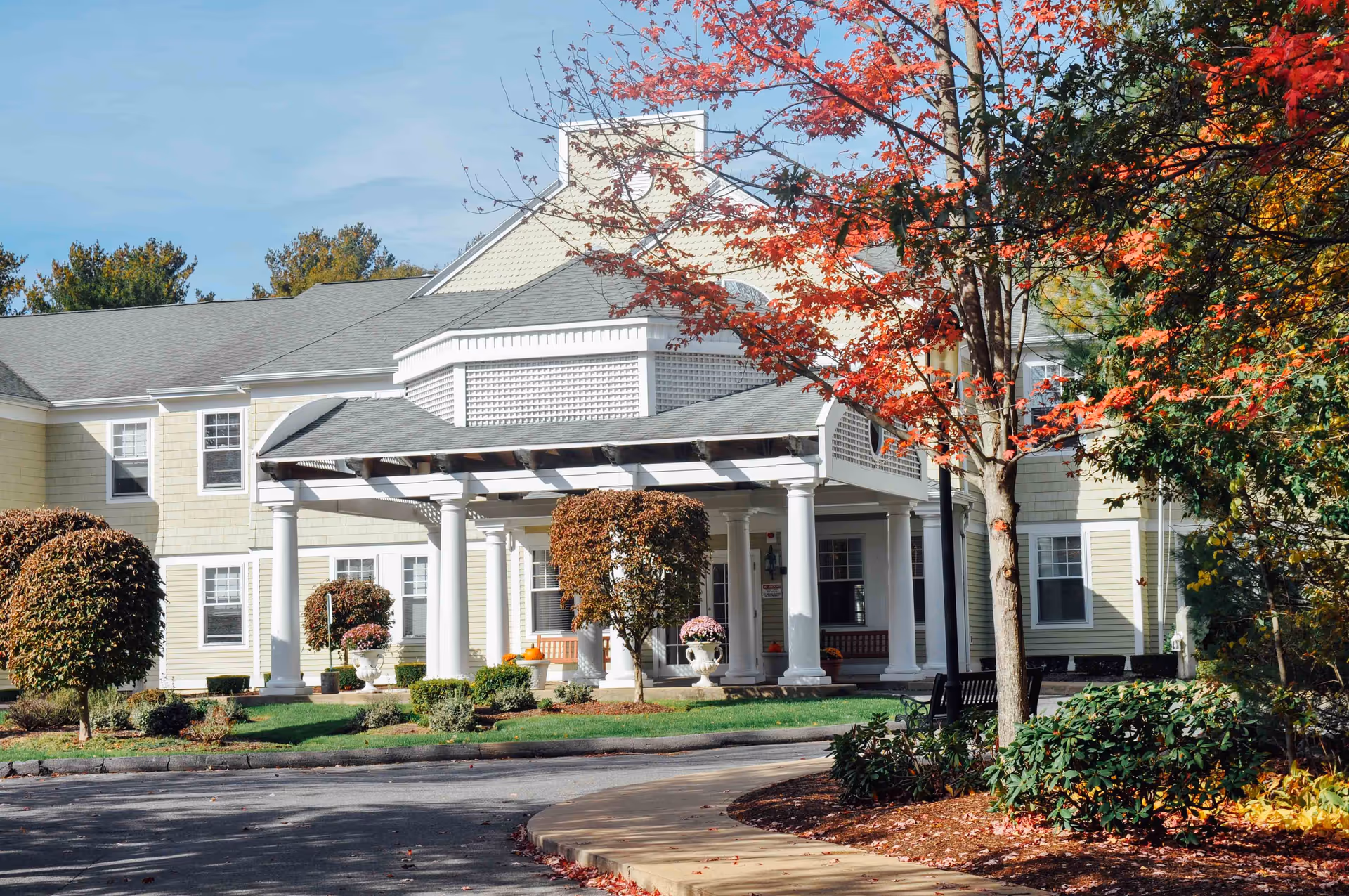 Exterior view of a senior living facility building with beige siding and white columns supporting a covered entrance. The scene includes well-maintained landscaping with trimmed bushes, a tree with red autumn leaves, and a curved driveway in front.