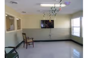 A small room with light yellow walls and a white tiled floor, featuring two chairs placed against the walls, a wall-mounted flat screen TV, and windows on two sides letting in natural light. Colorful streamers hang from the ceiling.