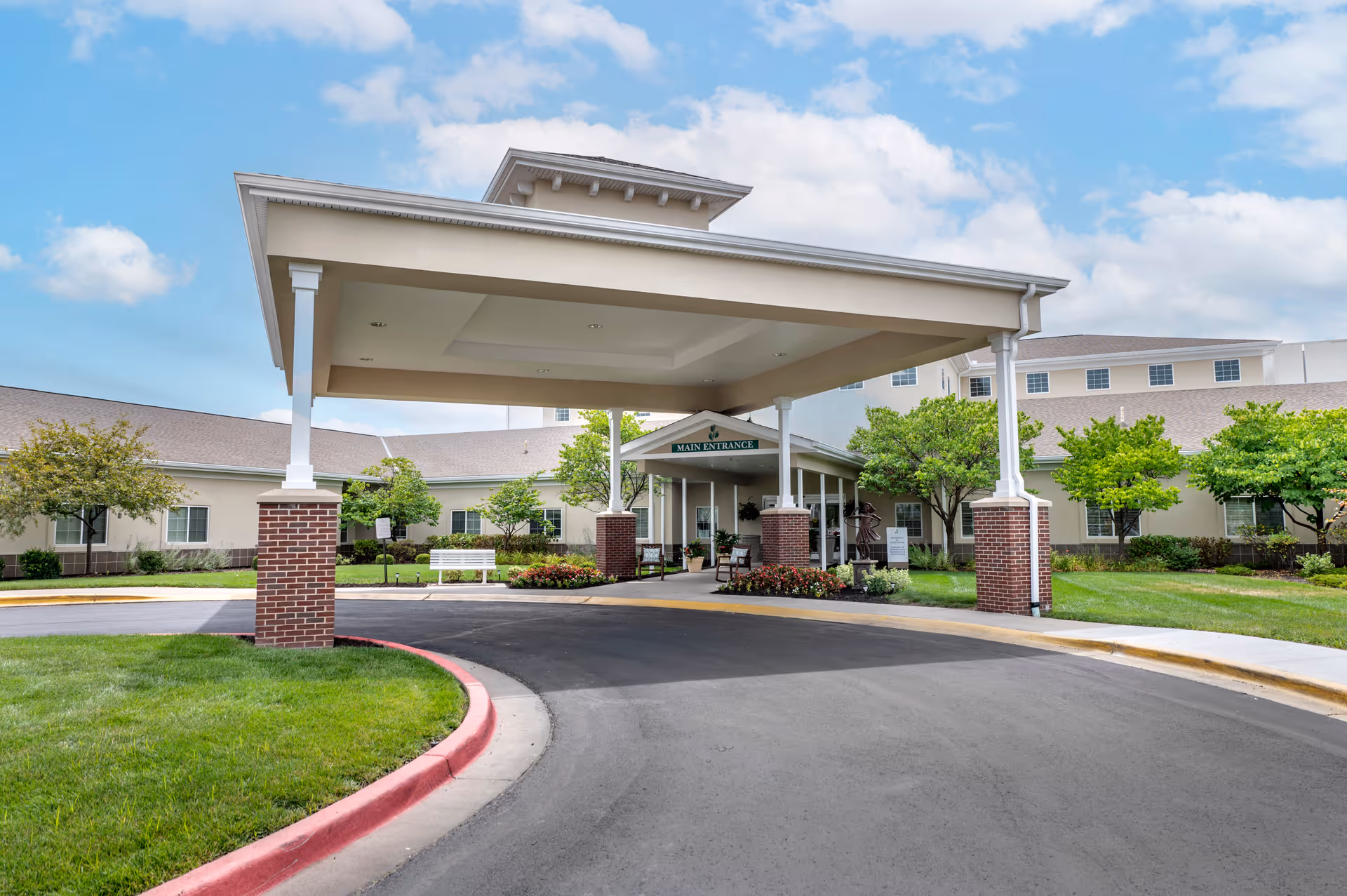 Front exterior view of Brookdale Overland Park senior living facility showing the main entrance with a covered drop-off area supported by white columns with brick bases, surrounded by green lawns, trees, and a clear blue sky with some clouds.