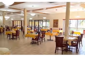 A spacious dining room with multiple wooden tables and chairs arranged neatly. The tables are covered with yellow tablecloths and set with plates and utensils. Large windows allow natural light to fill the room, and ceiling lights provide additional illumination. The room has a tiled floor and light-colored walls with some artwork hanging.