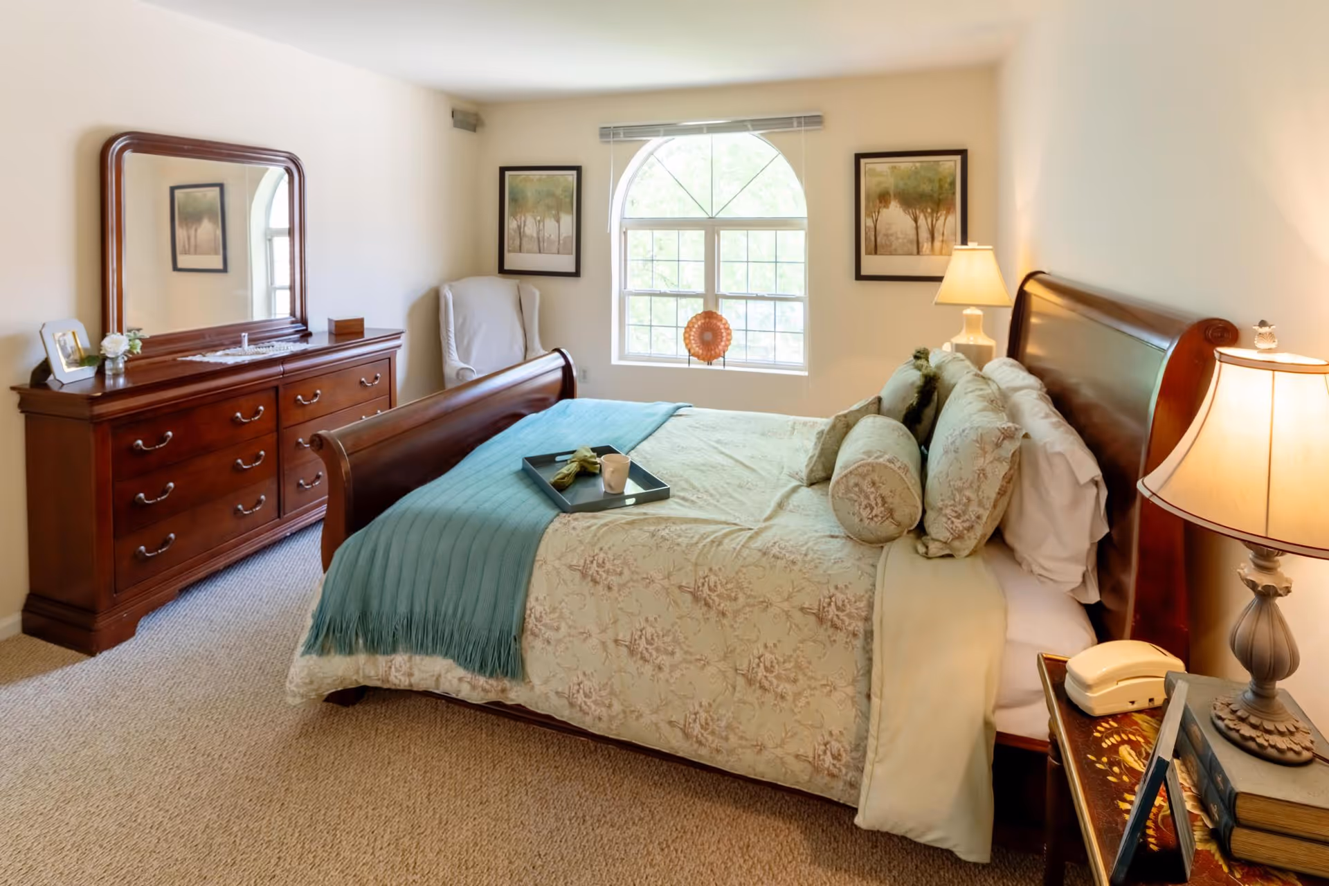 A cozy bedroom with a large wooden sleigh bed adorned with floral bedding and multiple pillows. A teal throw blanket is draped across the foot of the bed with a tray holding a cup and napkin. To the right of the bed is a nightstand with a lamp, books, and a vintage telephone. On the left side of the room is a wooden dresser with a large mirror and decorative items. A white armchair is positioned near a window with an arched top, flanked by two framed tree paintings. The room is softly lit and carpeted.
